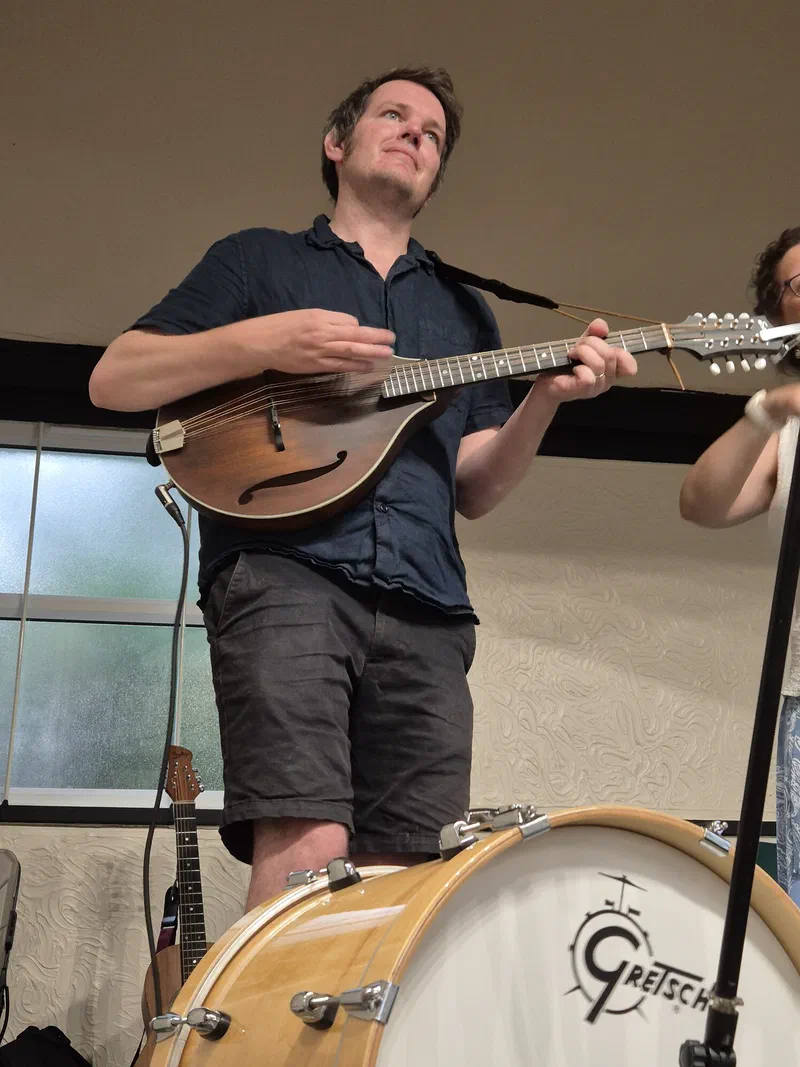 Tom playing the mandolin, seated by a drum kit