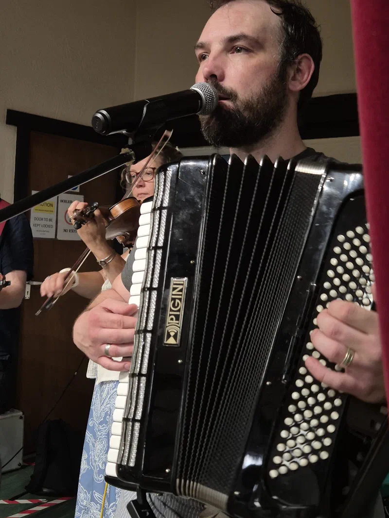 Dom singing into a microphone with his accordion, Sarah on fiddle behind him