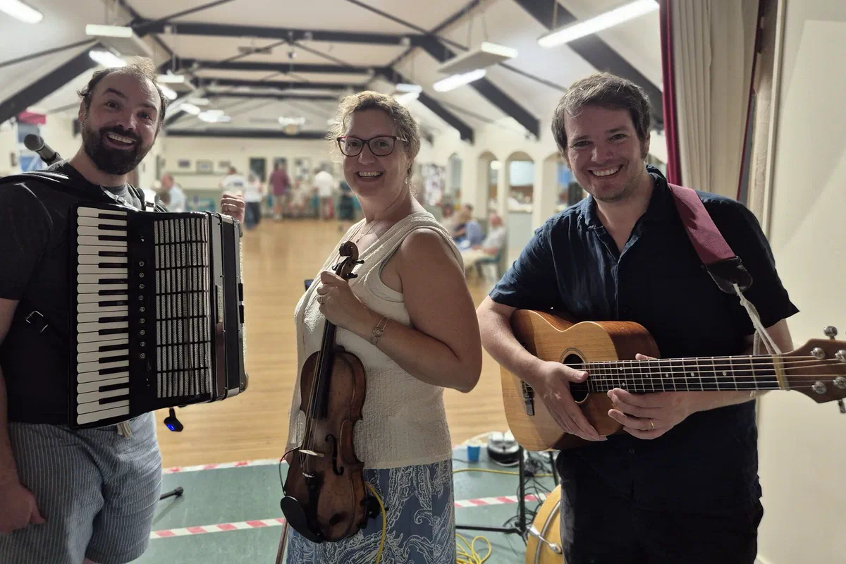 Dom, Sarah and Tom with accordion, fiddle and guitar in a village hall